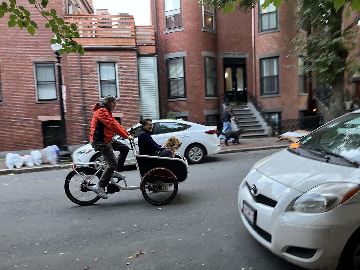 an older adult is giving a ride to a nother adult and a dog in an cargo e-bike