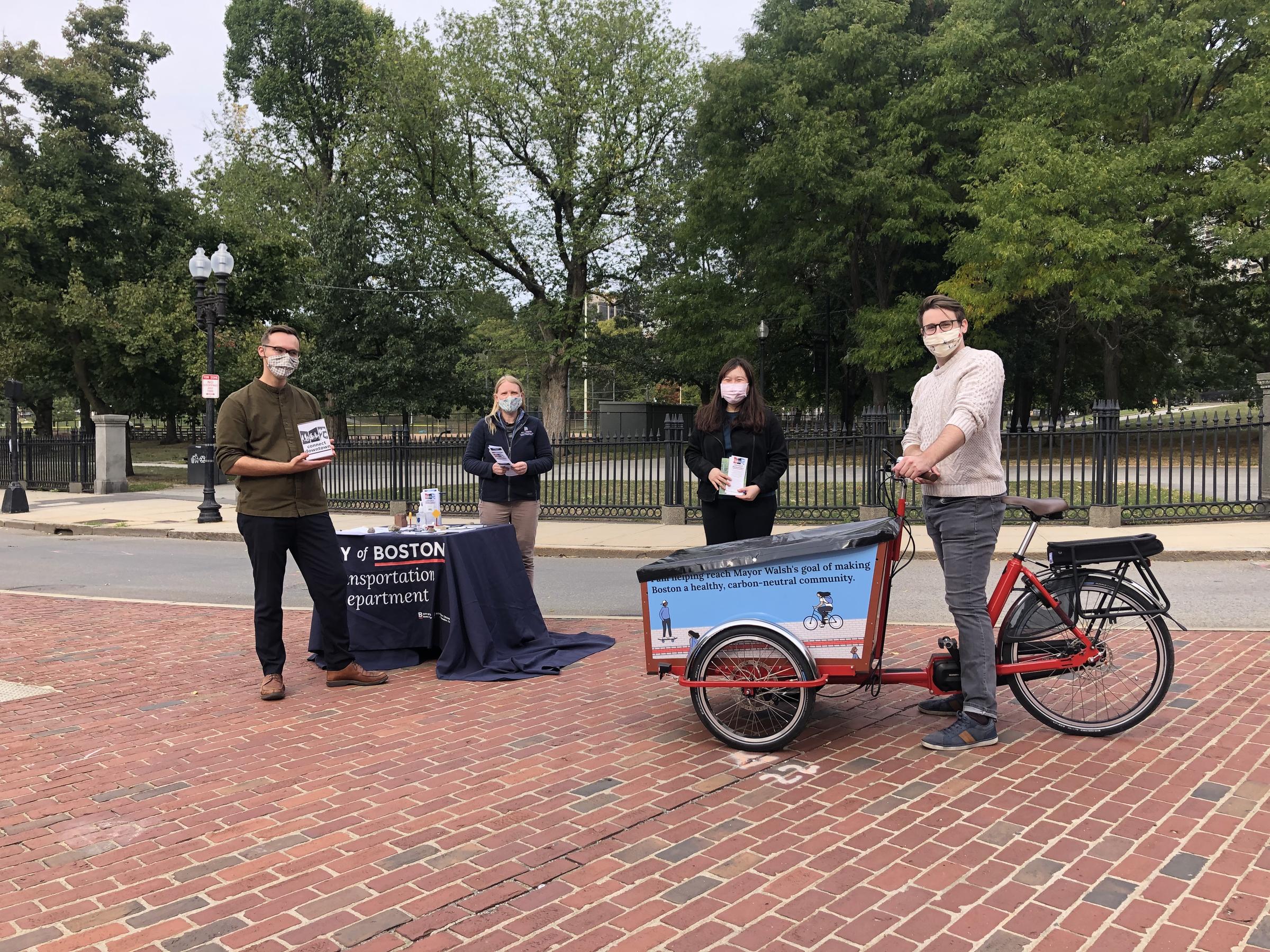 Four people wearing face coverings stand outside on a traffic island. Three people are holding brochures. A table has more materials and is covered with a blue tablecloth that says City of Boston Transportation department. One person stands near a cargo bike.