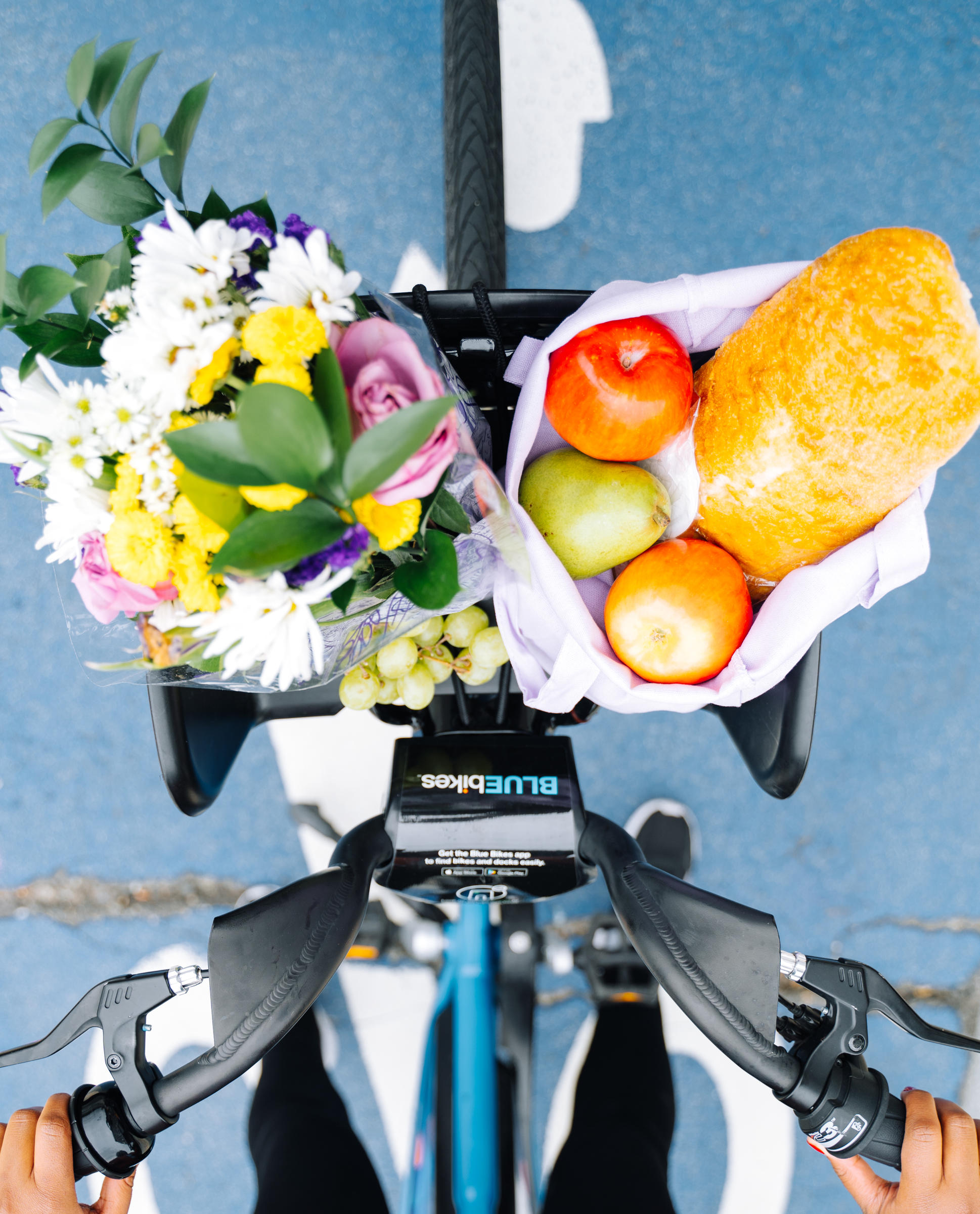 An overhead look at the front basket on a Bluebike. A bunch of flowers and a bag of groceries, including bread and apples, are in the basket.