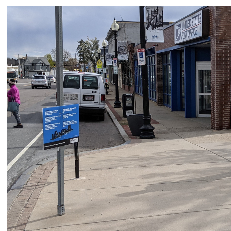 A blue sign located halfway up a pole.  The Hyde Park post office is to the right and the street is to the left.  The sign has information about potential Bluebikes station locations in the area