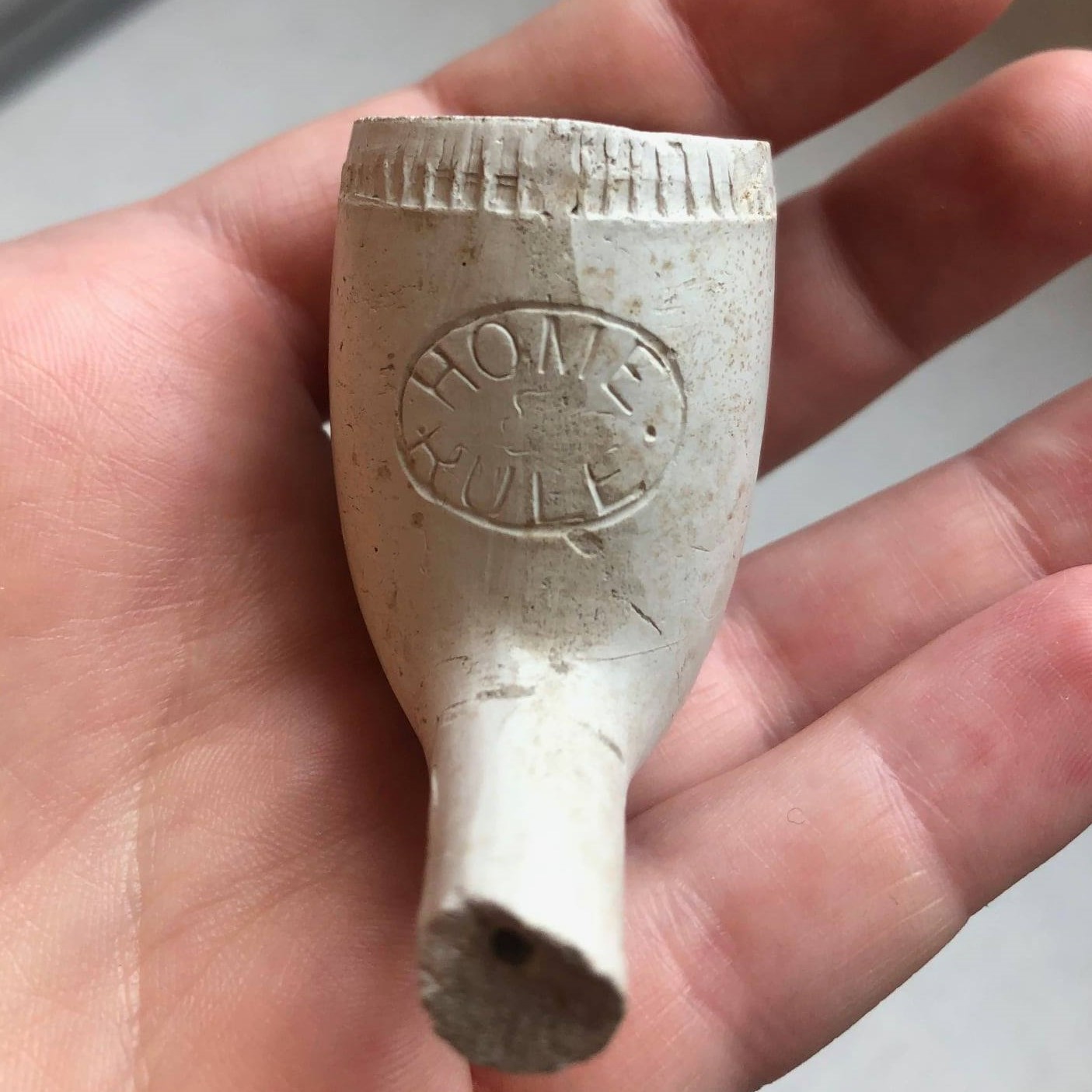 hand holding a fragment of white clay tobacco pipe with an impressed oval containing the words "HOME RULE" 