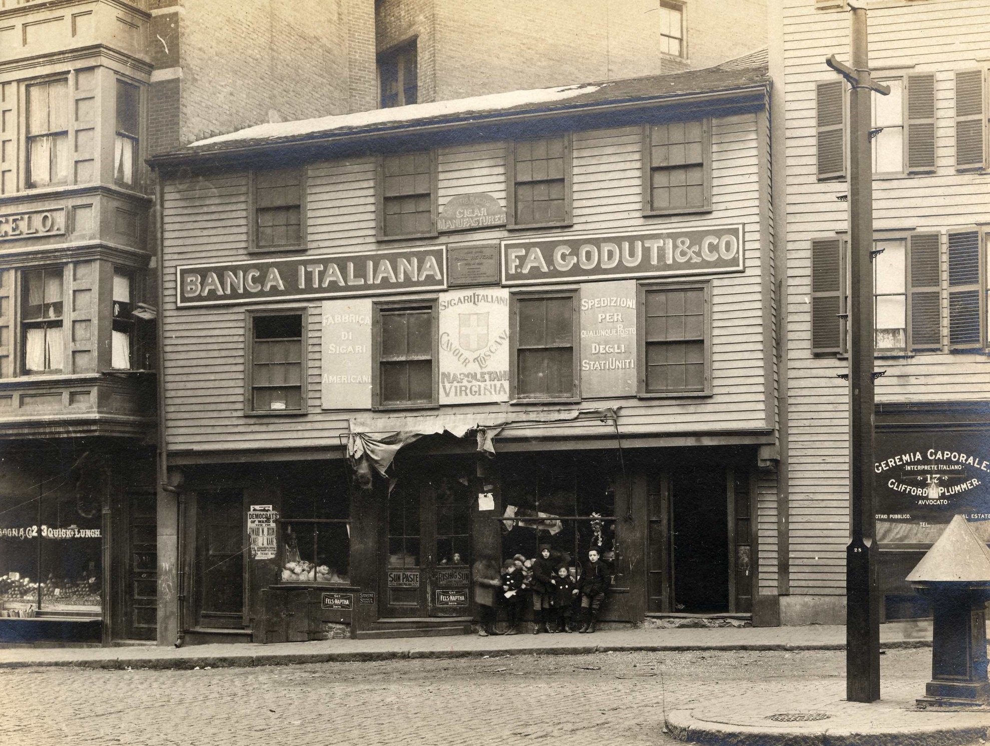Black and white image of a three story wooden building with windows on the second and third floor. First floor has store fronts with a group of children standing in front of them