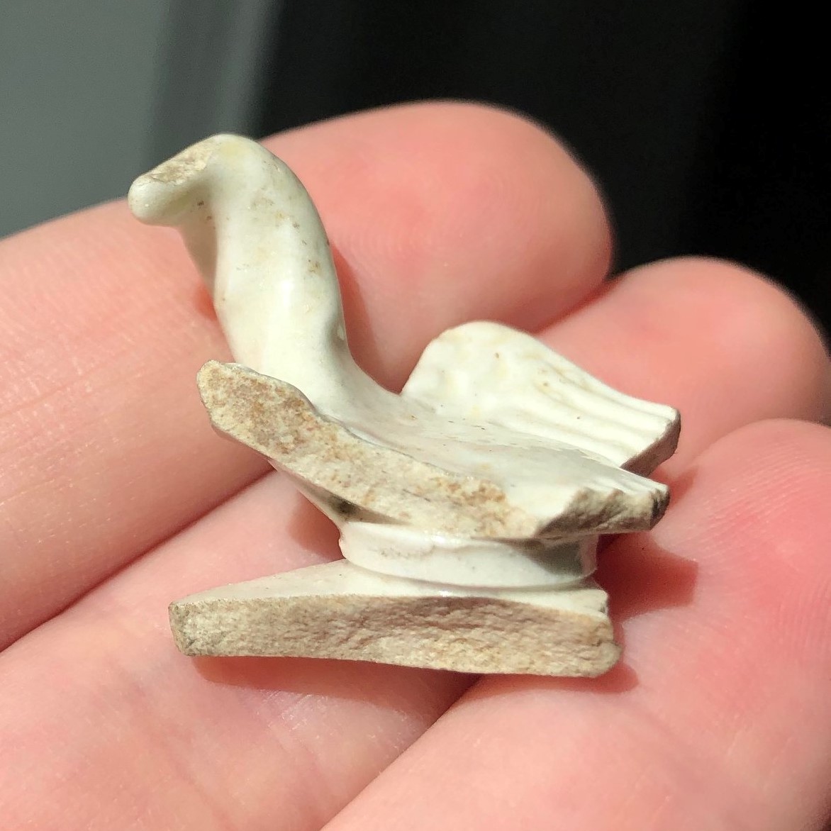fingers holding a fragment of white ceramic in the shape of a partially-broken bird with turned head and outstretched wings. Fragment is about one inch in all dimensions