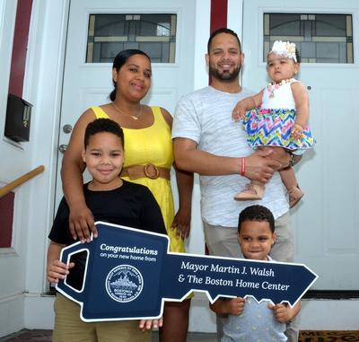 Image of family standing in front of housing, holding a symbolic "key" from the Boston Home Center.