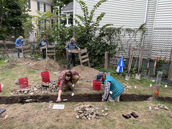 Photo of five women archaeologist working on a lawn. Three are standing at wood devices used to sift dirt on top of piles of dirt. Two are working at a long linear trench cut into the ground. There are buckets of dirt, shovels, and small piles of rock surrounding their work area.