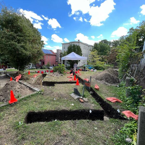 View of several open archaeological trenches in the front yard of an 18th-century structure on a sunny day. There are orange sandbags around the trenches and a white pop-up tent in the background