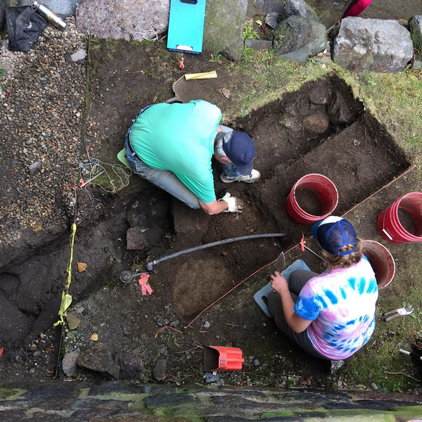 Two volunteers kneel beside a 1m x 3m archaeological unit with large cobbles and a round soil stain visible in the soil.