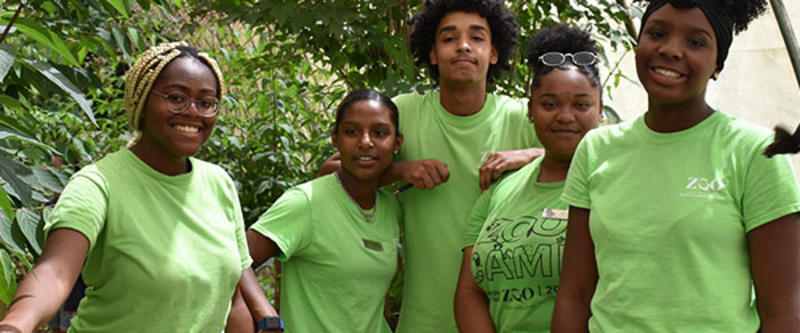 Image shows a group of young adults who are a part of the Youth Summer Jobs Program. They are all smiling and wearing light green shirts. 