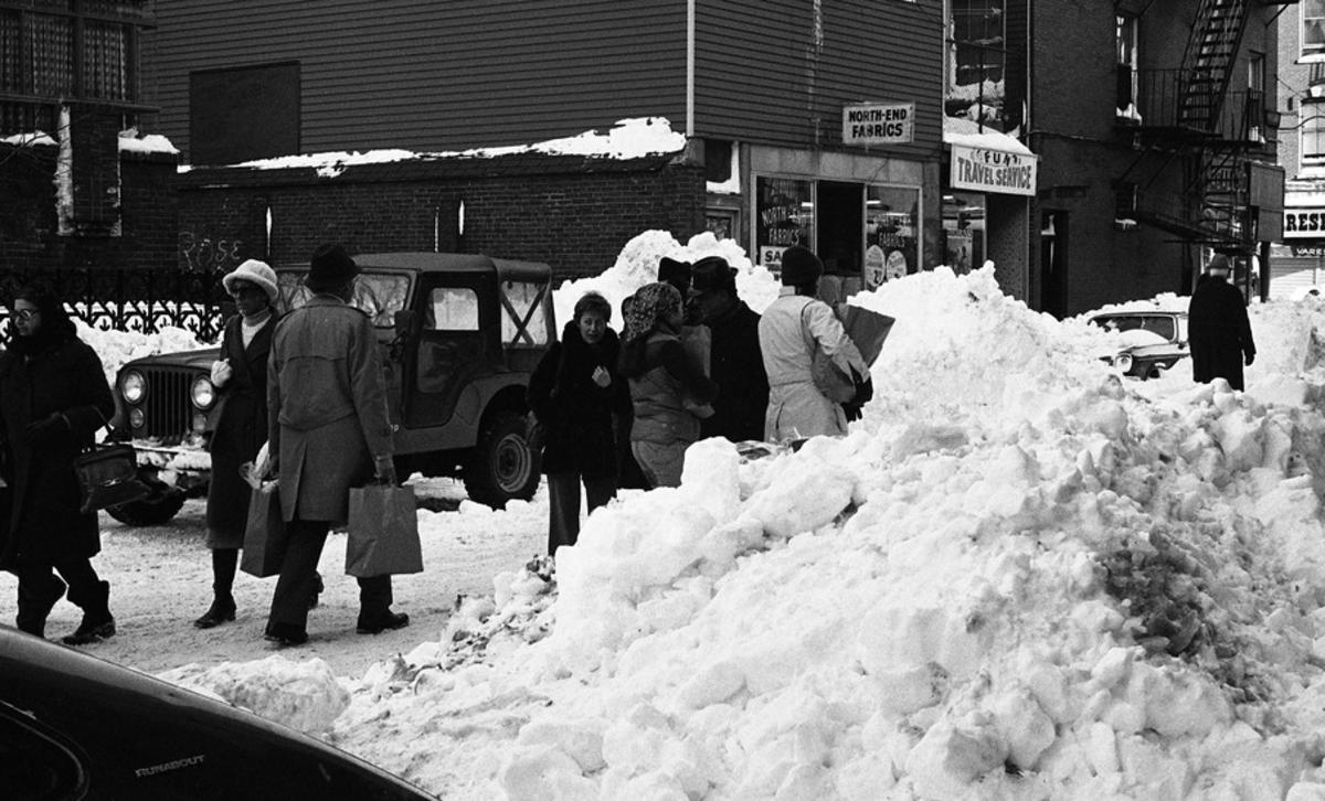Pedestrians carrying bags among snow piles on Parmenter Street