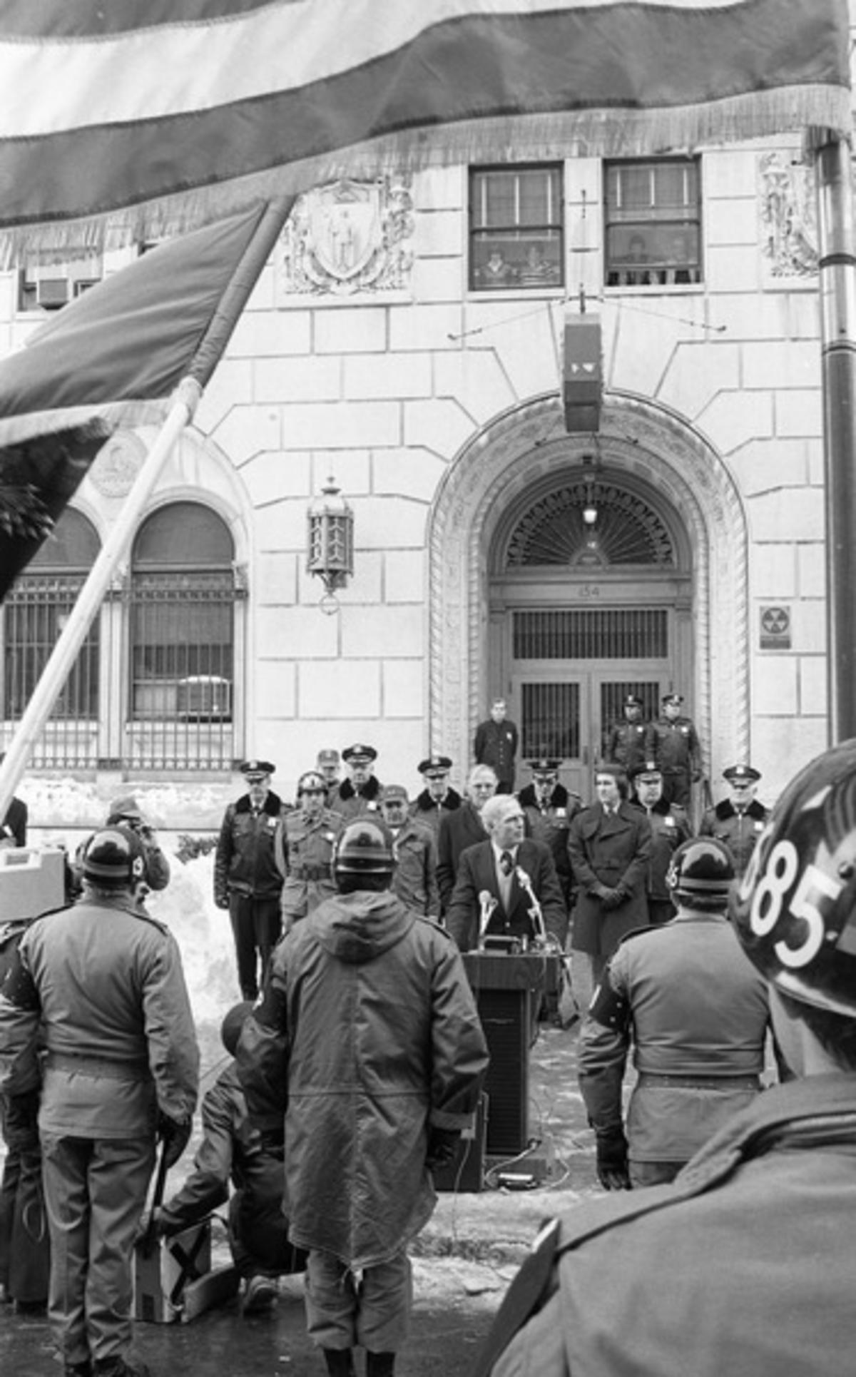 Mayor Kevin White standing at podium with police and Military Police Officers in front of Boston Police Headquarters