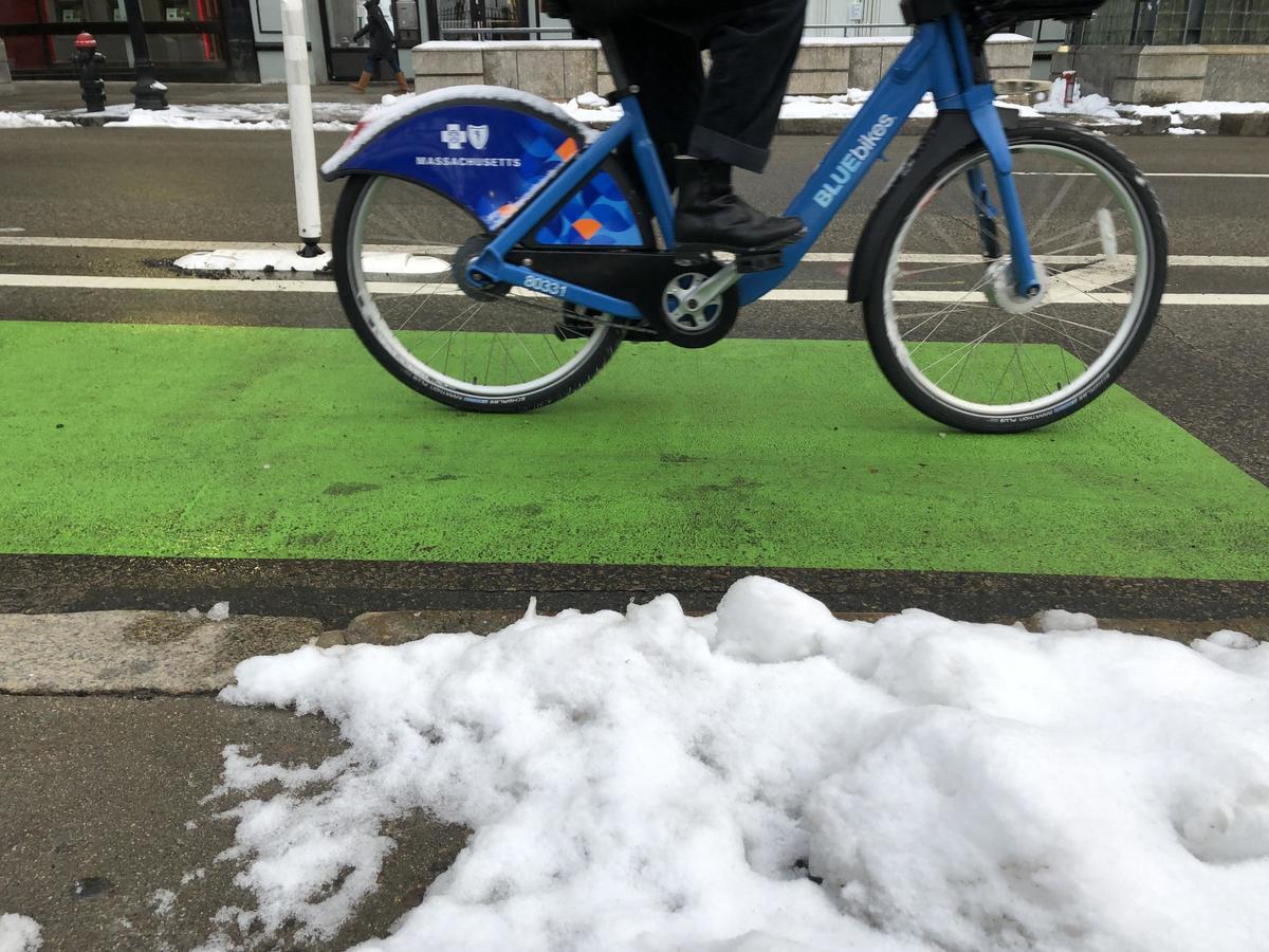 a rider wearing boots bikes on a bluebike in a bike lane painted green during the wintertime. Snow is present on the sidewalk