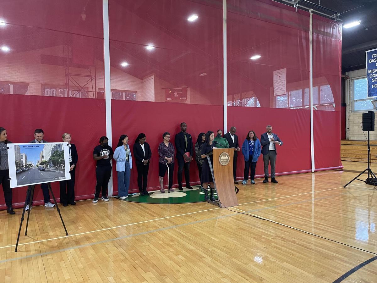 Mayor Michelle Wu in a school gymnasium delivering a press conference about bike network expansion; she is flanked by cabinet staff and community organizers.