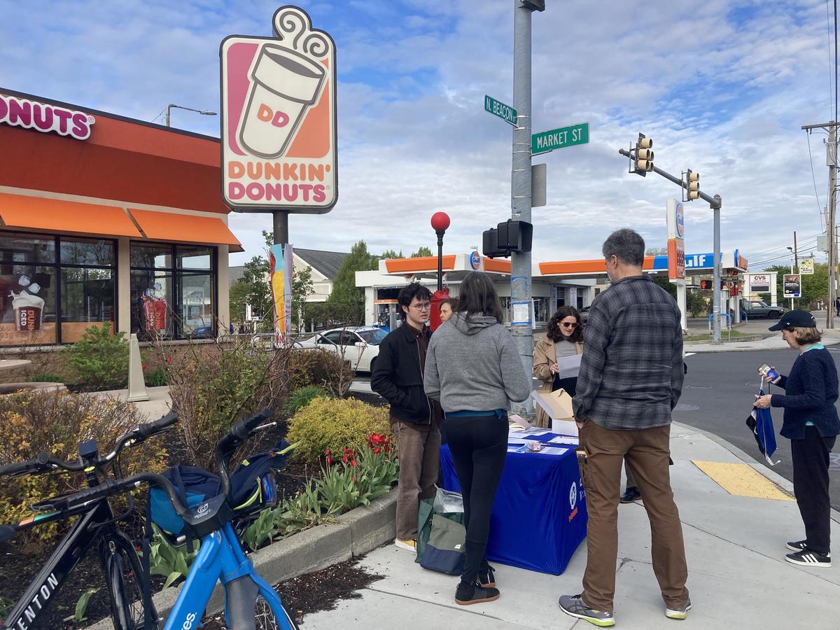 Group of five people standing around a table with flyers on it, at the corner of North Beacon St and Market St in front of the Dunkin Donuts