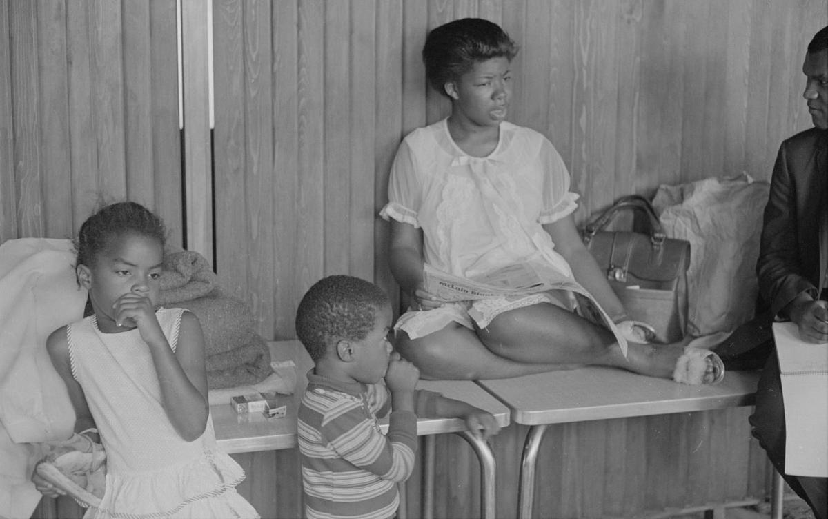 Black and white photo showing a mother sitting atop a table with her two children beside her. A reporter with a notebook is listening to her speak.