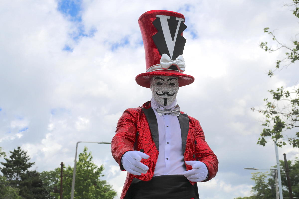 Street performer in a sparkly Guy Fawkes mask and red top hat with a large letter V on it.