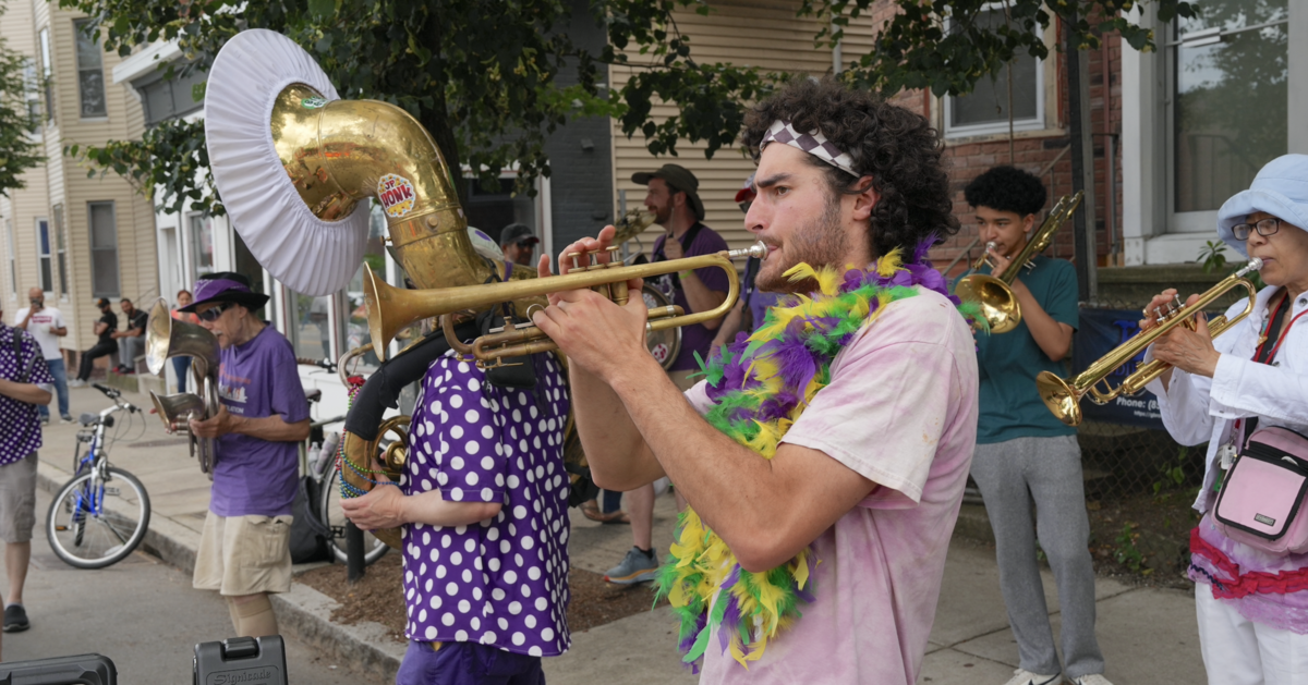 A band plays various brass instruments, in the foreground a man wearing a feather boa plays a trumpet