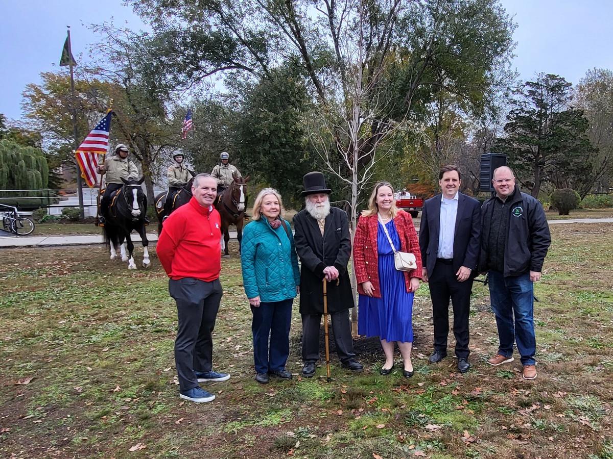 A group of six people stand around the newly planted tree. Three mounted rangers are in the background with a flag.
