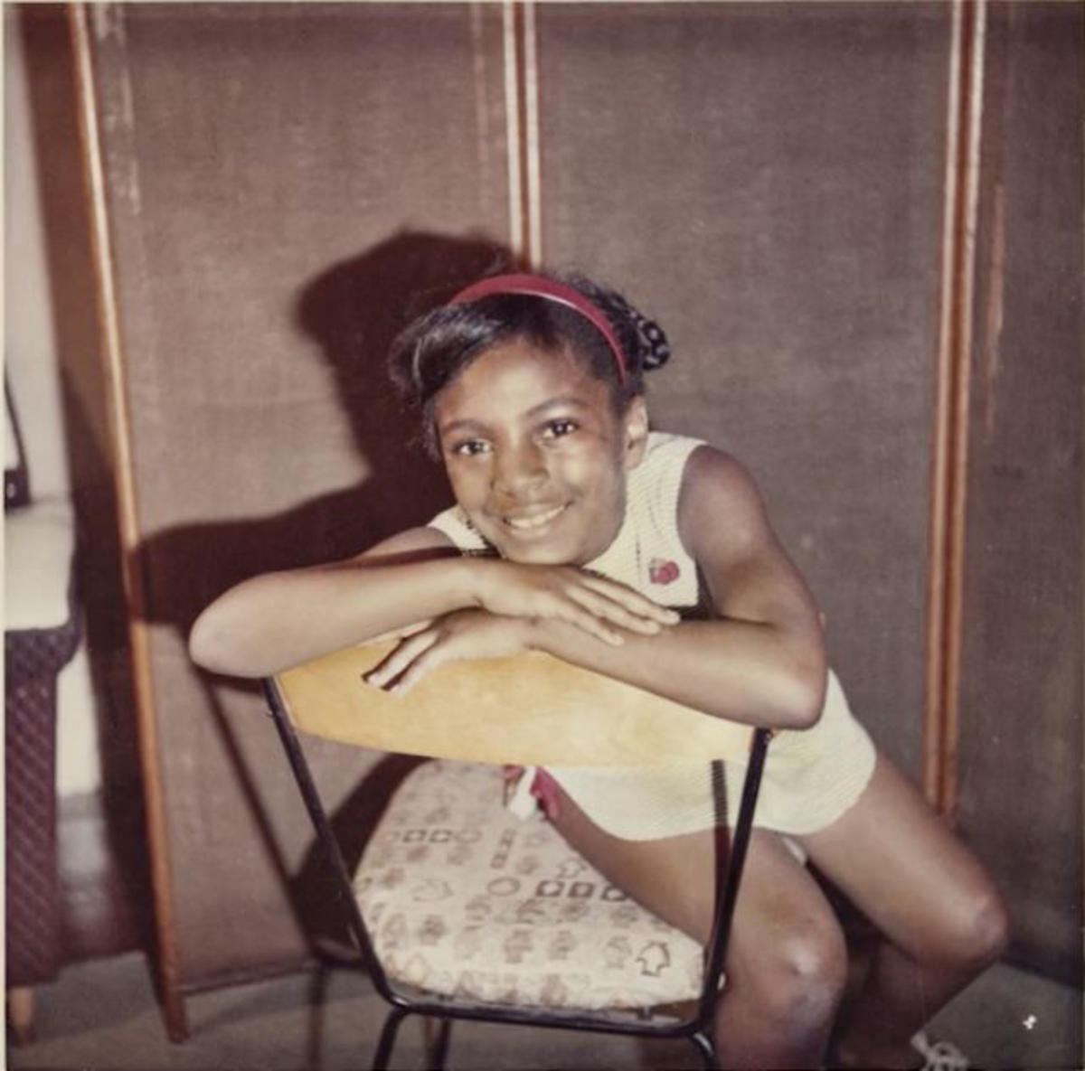 Image of Ekua Holmes as a young girl, sitting on a chair backwards with her arms crossed