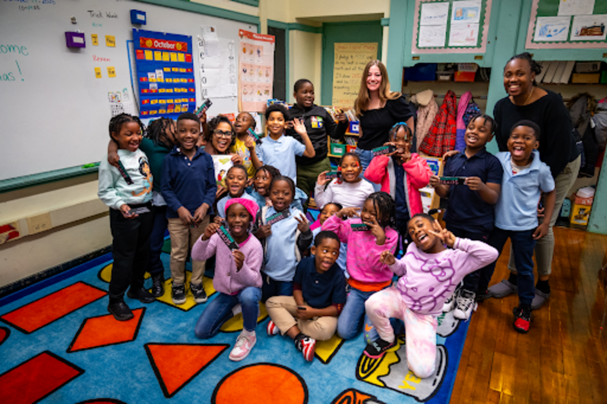 An image of children in a classroom with two adults standing behind them