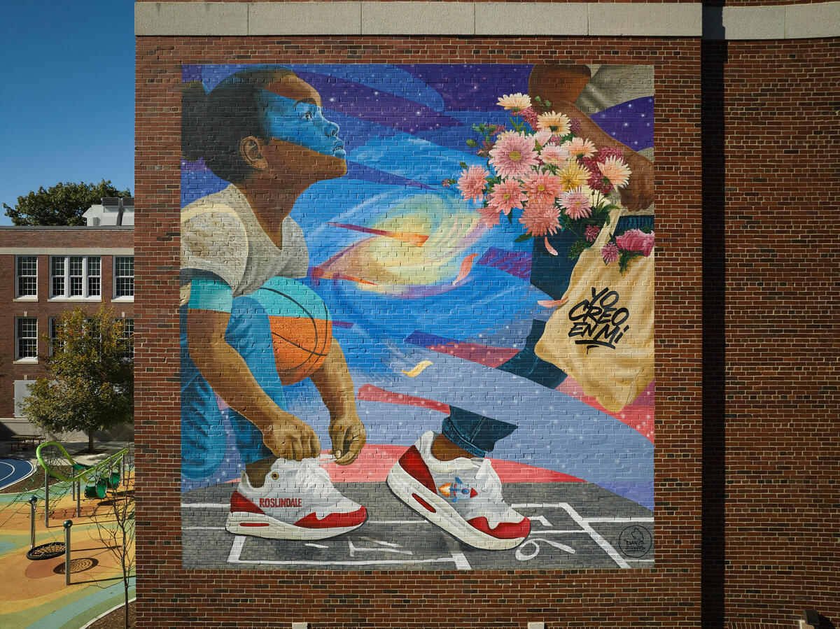 A young girl stops to tie her shoe, looking up at the person walking in front of her who carries a tote bag that says "Yo creo en mi"