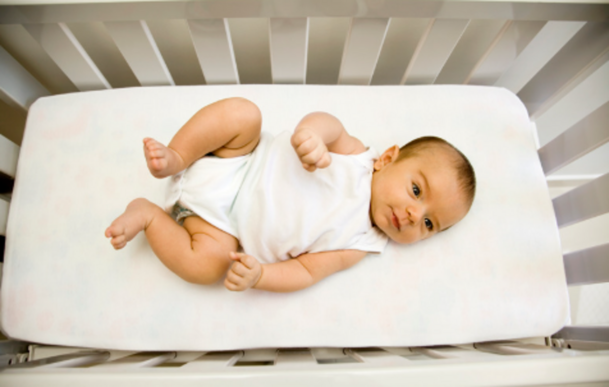 Baby in white onsie on their back in a crib. There are no toys, objects, or pillows and blankets surrounding the baby