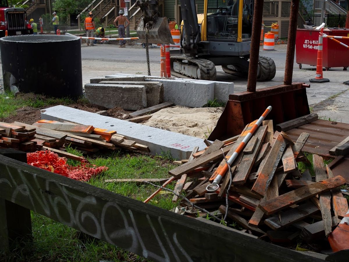 Piles of construction debris lie on a grassy median on the side of a road. Planks, blocks of granite, and large sewage pipes await installation, while construction crews stand in the distance.