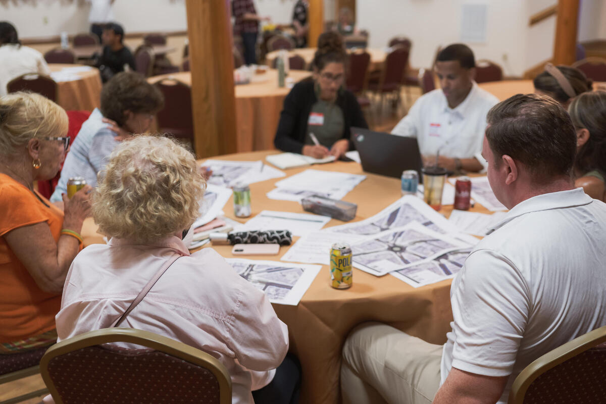 Eight people sitting around a circular table. One person takes notes on a laptop while another takes handwritten notes. Papers on the table show various concept designs for Andrew Square.