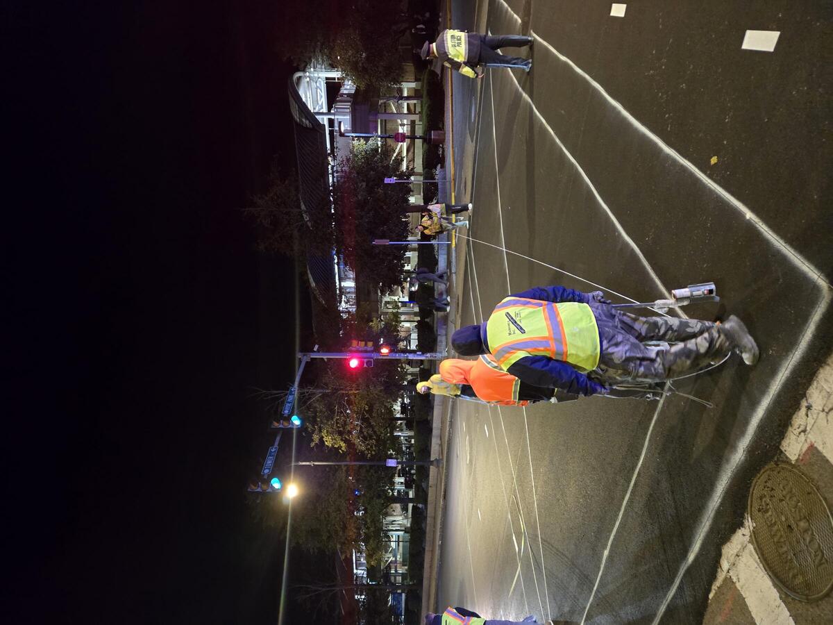 A photo showing a crew of men wearing fluorescent reflective vests installing markings on a freshly paved roadway at night in an urban environment.