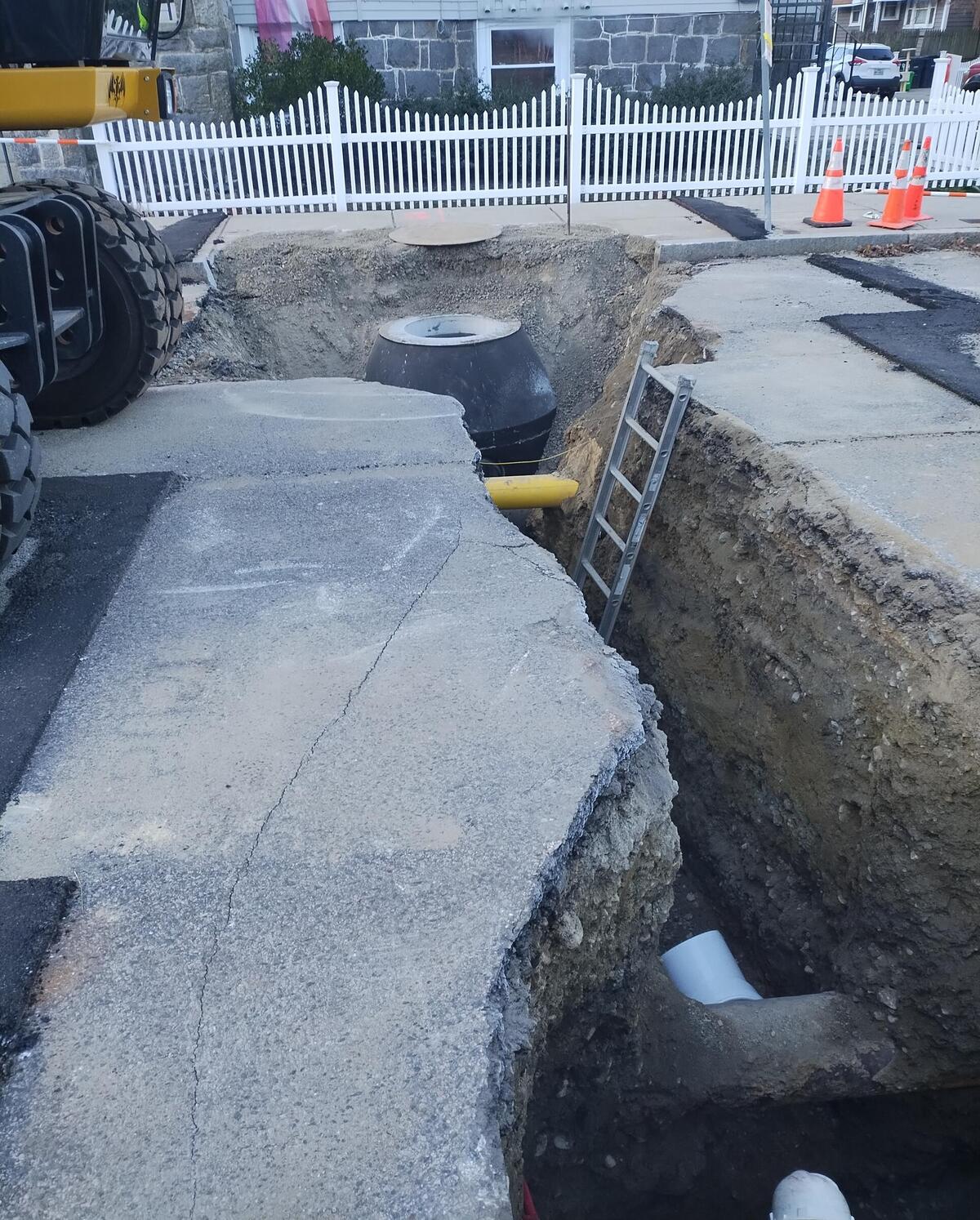 A street with a hole dug into the pavement and part of the sidewalk. Pipes and a structure can be seen exposed in the hole. A ladder extending into the hole and the back of a large truck are also visible.
