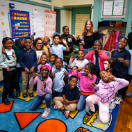 An image of children in a classroom with two adults standing behind them