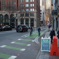 On State Street, two bicyclists travel in a bike lane toward the Old State House. Cars are parked along the left curb, and one car is being driven on the street. Pedestrians in winter coats enter a crosswalk.