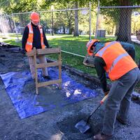 Two archaeologists in blaze orange safety vests and hard hats excavate an archaeological test pit on Boston Common.