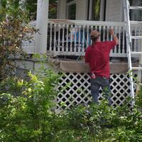 A man in a red shirt paints a white porch while next to a ladder