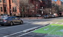 A gray sedan drives on a Boston street beside a green separated bike lane. The bike lane is delineated by concrete barriers and white markings. Parked vehicles line the far side of the bike lane, and brick buildings form the streetscape under a sunny sky.